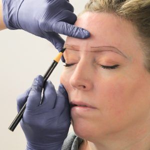 Hands holding black pencil against woman's eyebrows, drawing eyebrow shape onto skin.