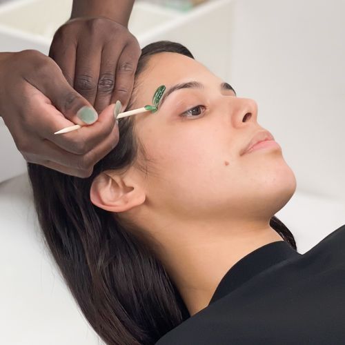 Hands applying green facial wax to skin above brunette woman's eyebrow arch.