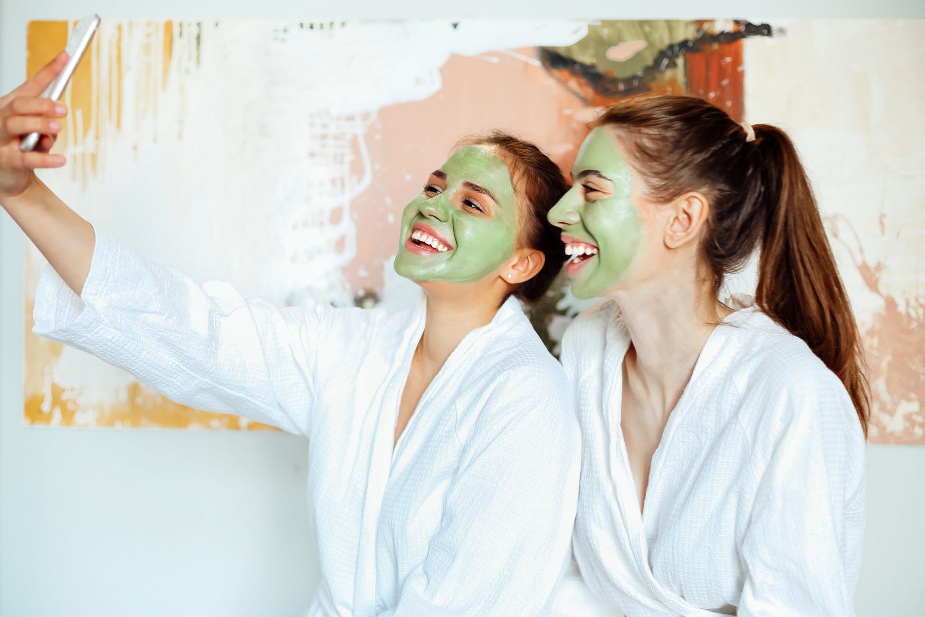 Two women in spa robes with green face masks taking a selfie, highlighting fun and relatable moments in social media for estheticians