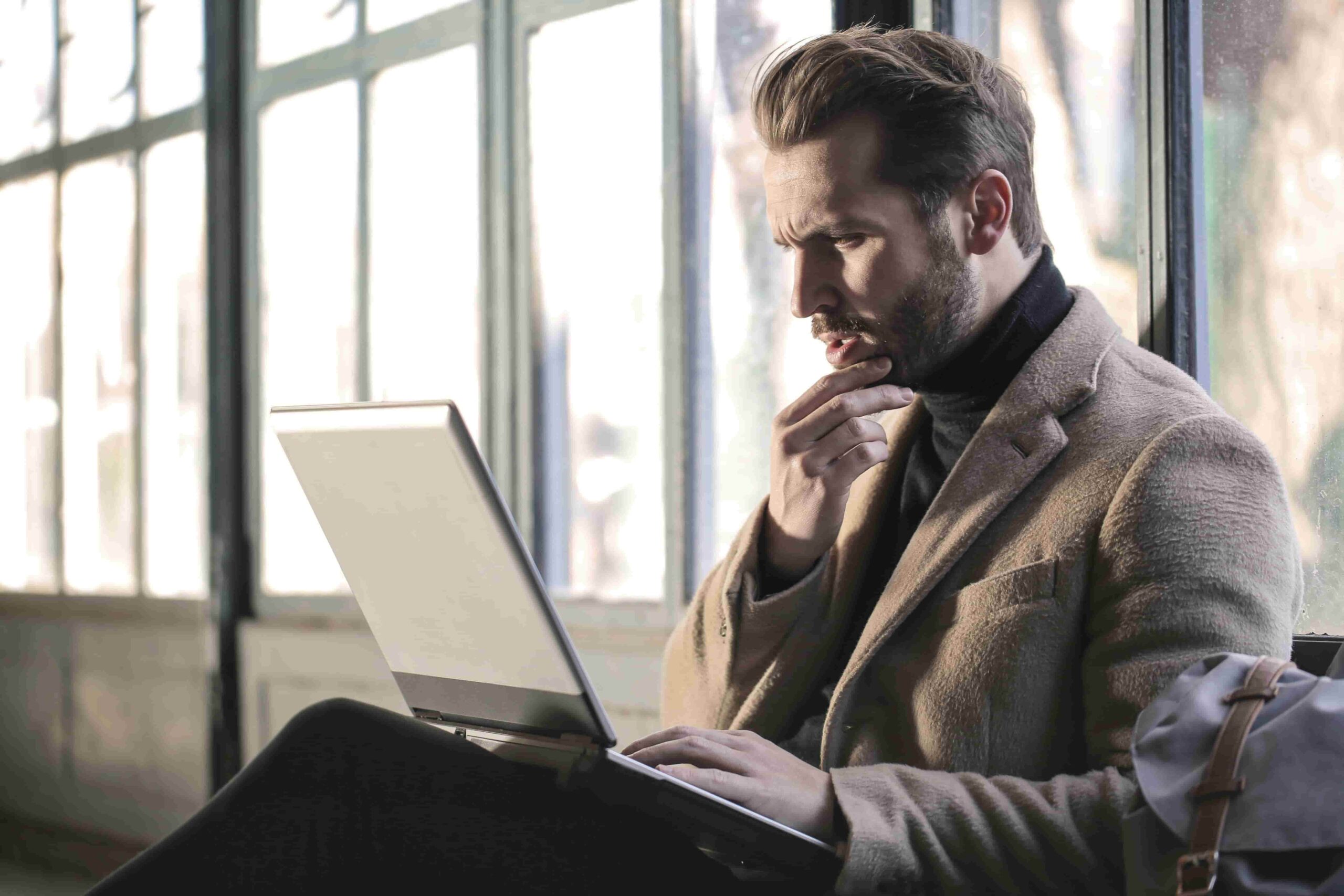  A man sits by a window with a laptop on his knees, leaning forward with his hand on his chin in focused curiosity. The image illustrates the interested phase of The Client Decision Funnel, where a potential client is actively engaging and seeking more information.