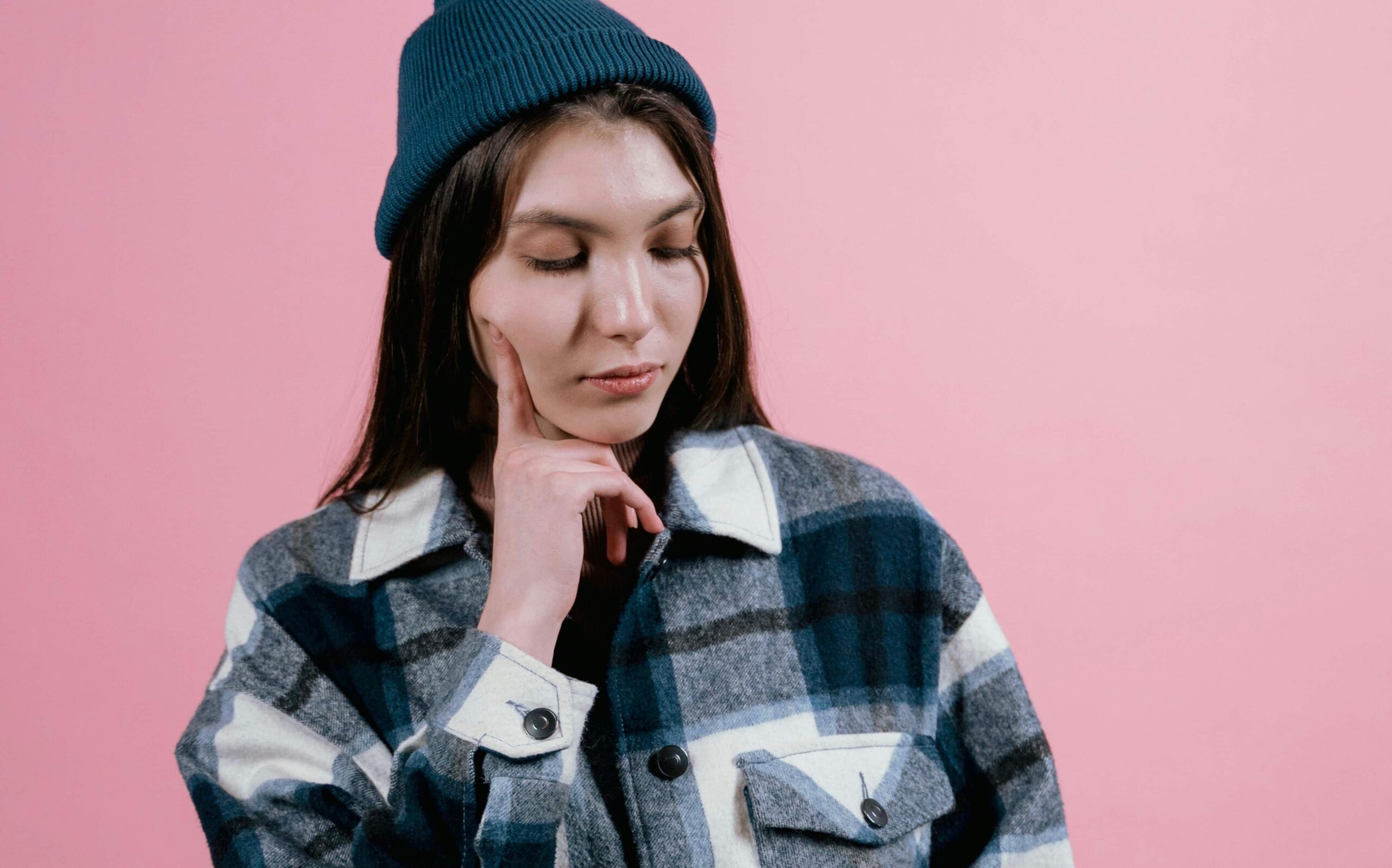 A young woman wearing a beanie and plaid jacket rests her finger on her cheek, looking down thoughtfully against a pink background. The image illustrates the consideration phase of The Client Decision Funnel, symbolizing a client weighing options before making a decision.
