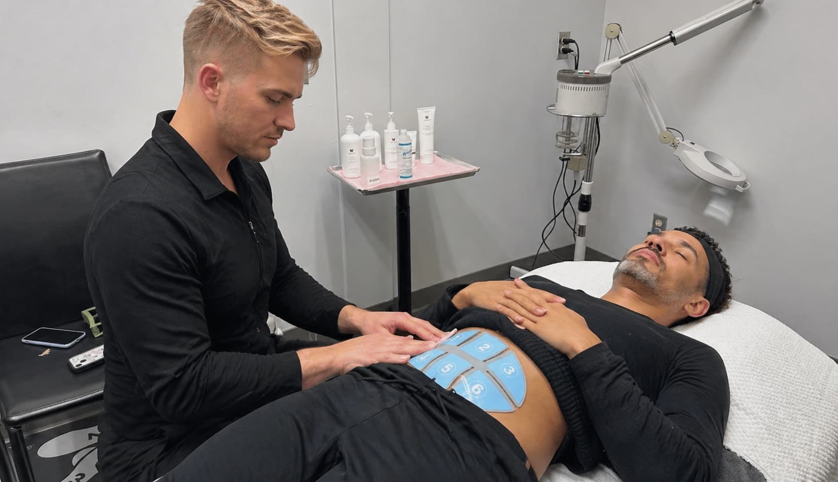 A hands-on training moment from a laser technician course Winnipeg set in a clean, clinical treatment room, where a practitioner adjusts blue body contouring pads on a client’s abdomen. The client lies relaxed on a treatment bed with hands resting on their chest, wearing a headband and comfortable clothing. The practitioner sits beside the bed, focused on proper placement and technique. In the background, a magnifying lamp, organized skincare products on a tray, and minimal medical equipment reinforce a professional learning environment centered on advanced aesthetic treatments.