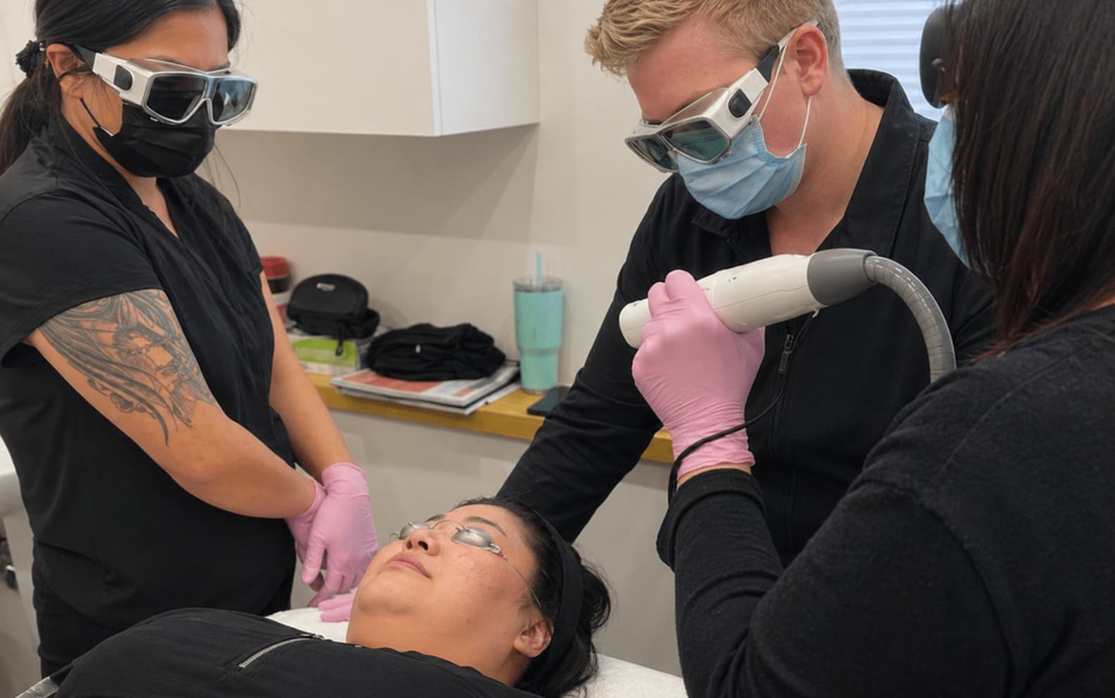 A hands-on training session from a laser technician course Winnipeg in a bright clinical treatment room, where a practitioner uses a handheld laser device on a client’s cheek while the client lies on a treatment bed wearing protective eye shields. Two additional practitioners in black attire and protective eyewear observe closely, focusing on technique and positioning. One practitioner stands near the client’s head while another stands to the side, creating a collaborative learning environment. The space features neutral walls, organized tools and supplies on a counter, and professional equipment, reinforcing a clean, structured setting for advanced laser training.