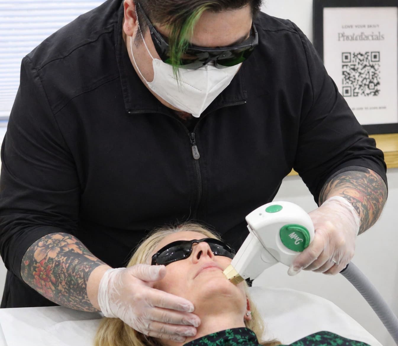 Close-up of a hands-on training moment from a laser technician course Winnipeg, showing a practitioner wearing protective eyewear, a face mask, and gloves performing a facial laser treatment on a client lying on a treatment bed. The client wears dark protective goggles and rests comfortably with a headband in place while the practitioner uses a handheld laser device on the lower face. The practitioner’s forearms, visible with tattoos, and a small section of hair with a coloured strand can be seen as they focus on precise technique. The clean, clinical room features soft lighting, neutral walls, and framed educational materials in the background, creating a professional environment for advanced laser training.