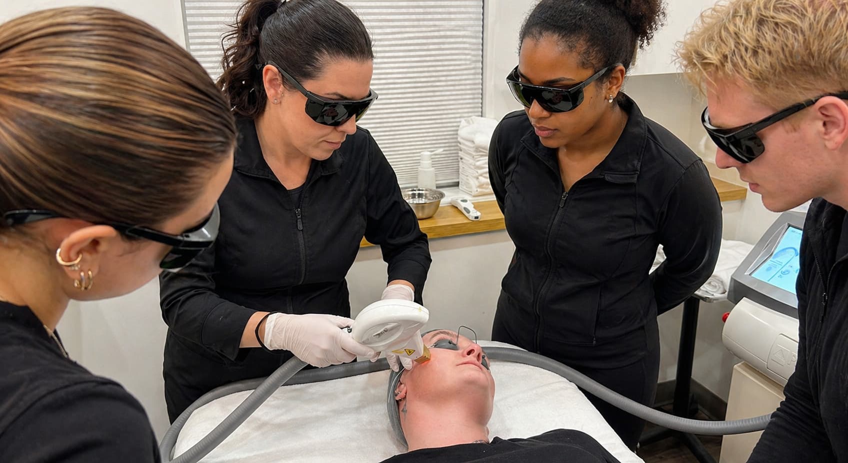 A hands-on training session from a laser technician course Winnipeg in a bright clinical treatment room, where a practitioner uses a handheld laser device on a client’s cheek while the client lies on a treatment bed wearing protective eye shields. Three observers in black attire and safety eyewear stand closely around, watching the technique with focused attention. The space includes neatly arranged towels, skincare supplies on a counter, and professional equipment nearby, creating a clean, structured learning environment centered on safe and precise laser application.