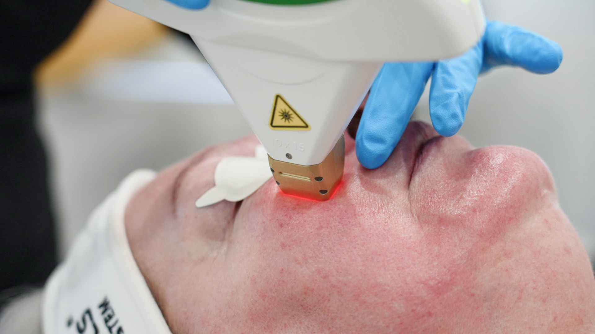 Female esthetician wearing black professional smock and white face mask using hand to gesture towards laser hair removal machine.